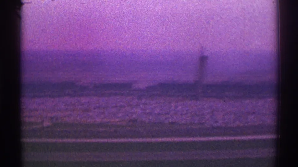 Purple-tinted 8mm frame from a 1969 Los Angeles airplane view, showing open land, distant low buildings, haze, and a tower form.