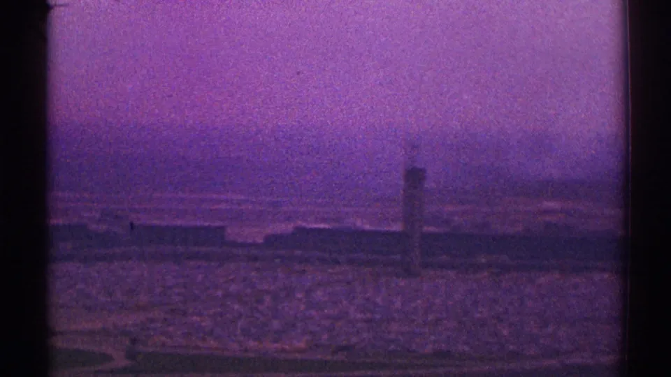 Late-1960s Los Angeles aerial home-movie frame with open ground, distant urban forms, and a tower-like structure softened by haze and film grain.