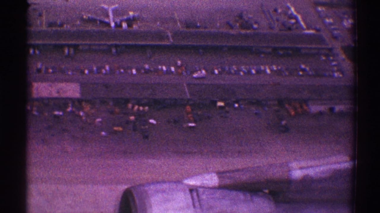 Jet engine and wing seen over airport buildings and parked cars during takeoff.