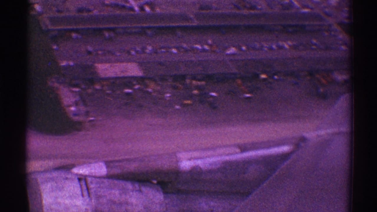 Aircraft wing and engine over airport pavement, buildings, and a line of parked cars.