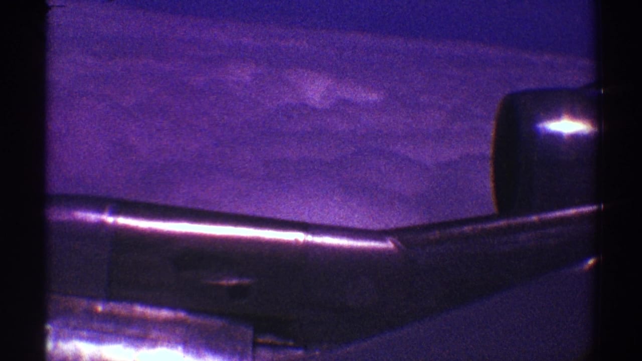1969 8mm-style view from an airplane window over Los Angeles, showing a dark wing against cloud cover and blue-purple sky.