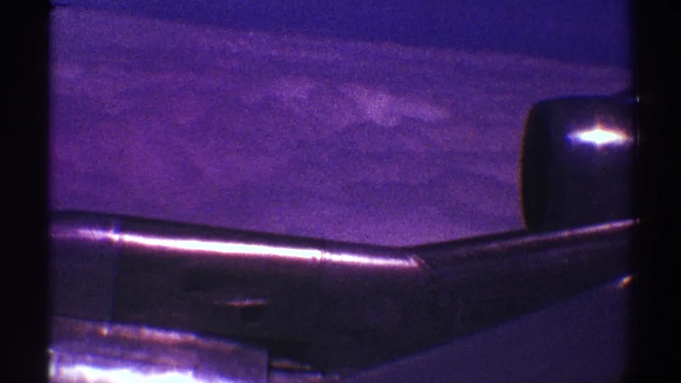 Aerial passenger view of a dark aircraft wing above clouds, with heavy grain and a purple color shift from the 8mm scan.