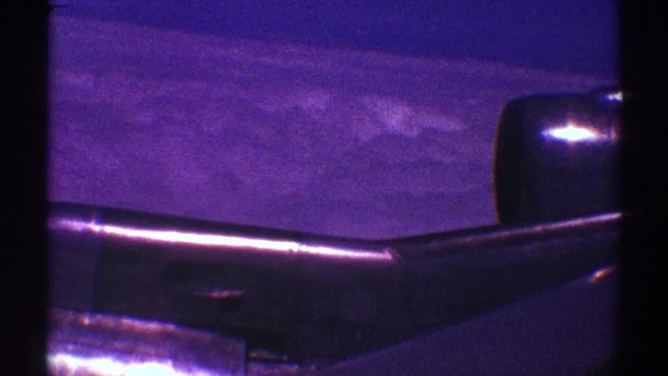 Passenger-seat aerial view showing the aircraft wing low in frame, cloud layers beyond, and visible 8mm grain and magenta cast.