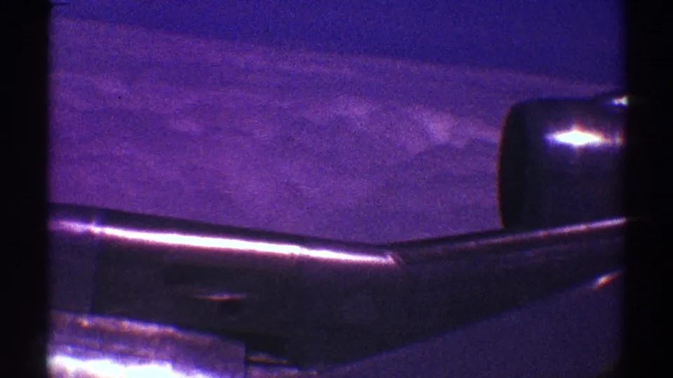 Late-1960s home-movie frame of an airplane wing and rounded engine area over cloud cover, with dark borders and purple-blue film color.