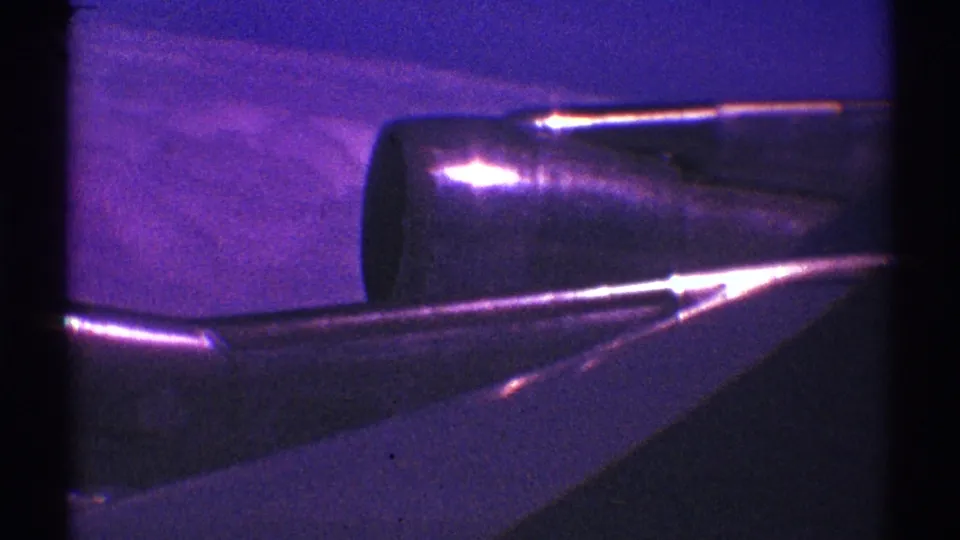 Purple-tinted 8mm frame showing an airplane engine beside the wing, with soft sky or terrain beyond.