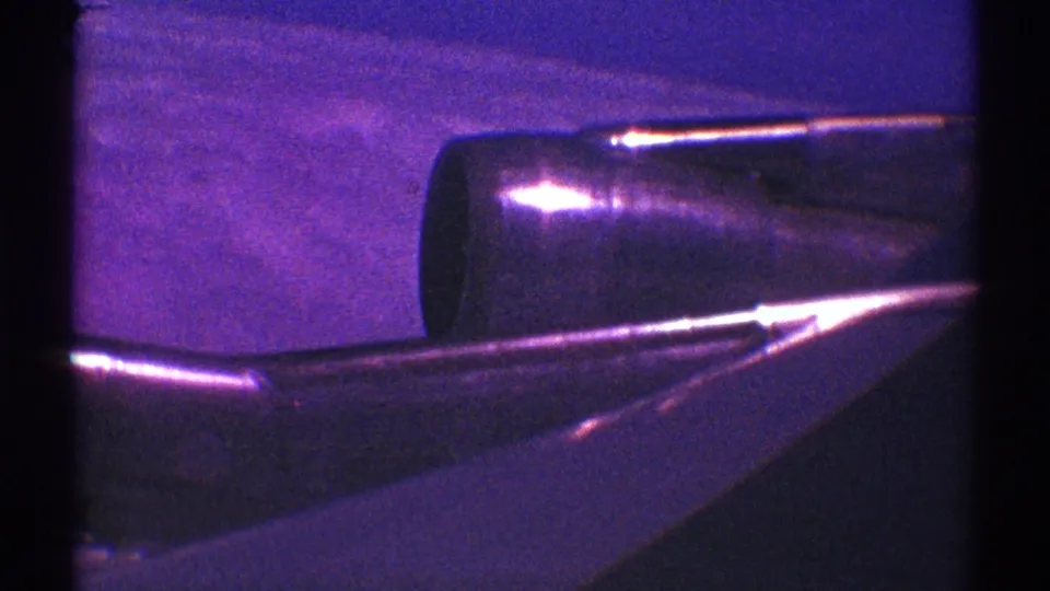 1969 home-movie frame of a wing and engine in flight, with heavy grain, purple cast, and reflected light.