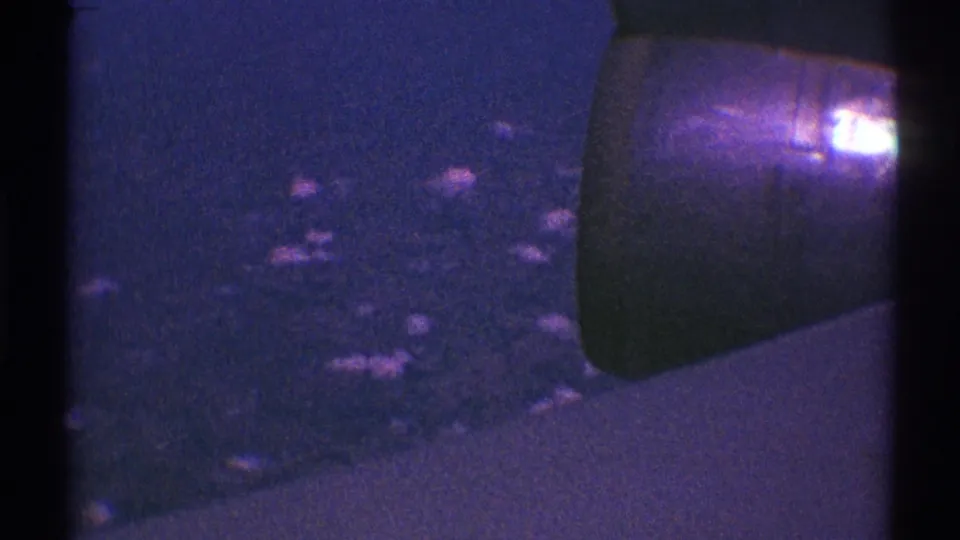 Purple-blue 8mm aerial view from a passenger window, with the engine at right and indistinct land or clouds below.