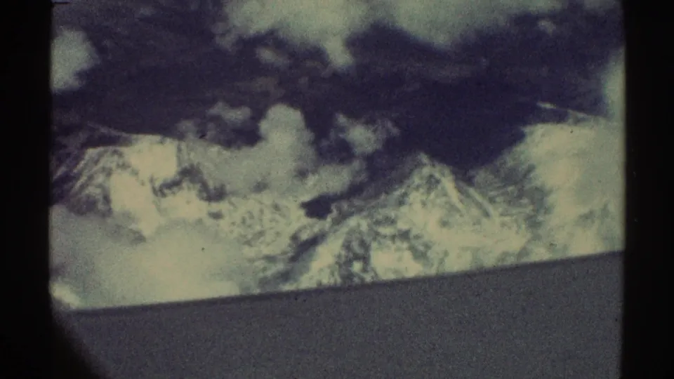 1969 8mm frame with snow-covered peaks, low cloud shadows, and a muted gray foreground edge from an apparent airplane view.