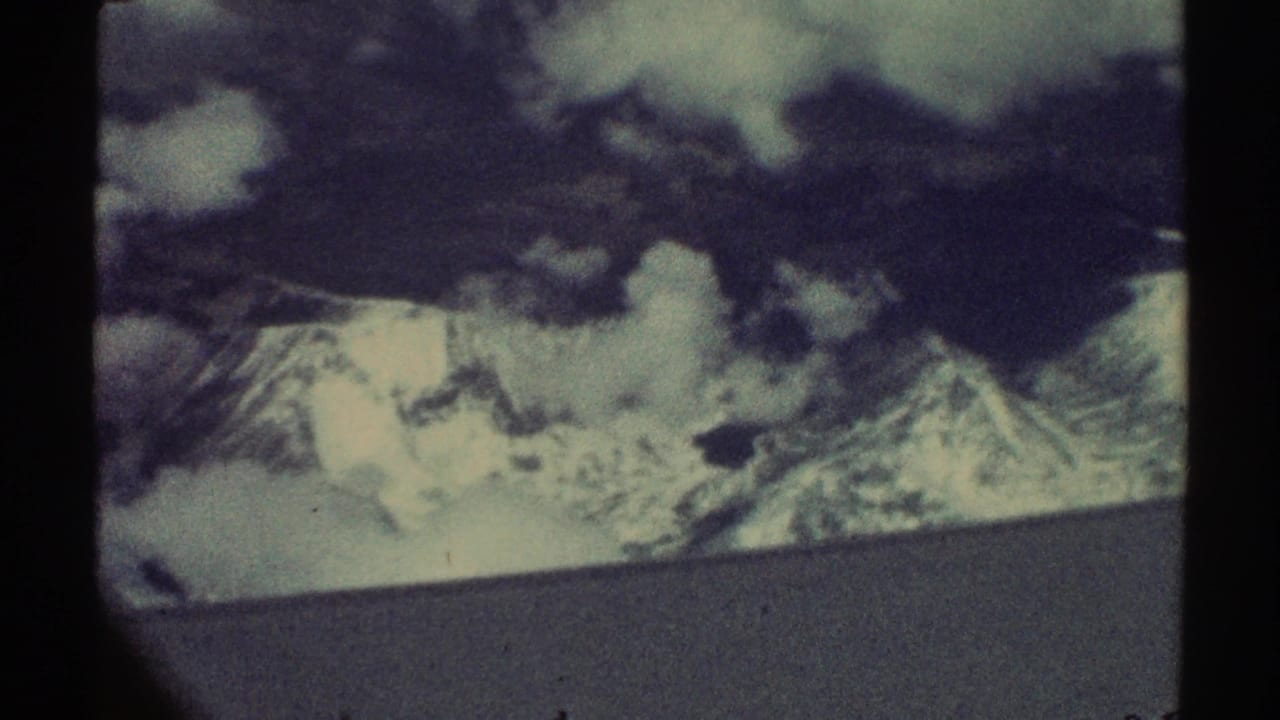 1969 8mm home-movie frame showing distant snow-capped mountains and clouds from an airplane-window perspective.