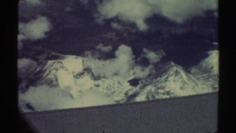 1969 8mm home-movie frame showing distant snow-capped mountains and clouds from an airplane-window perspective.