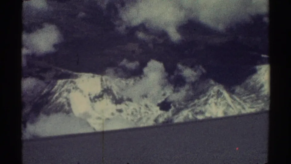 Vintage 8mm frame of snowy mountain ridges partly obscured by clouds, filmed from an aircraft in 1969 travel footage.