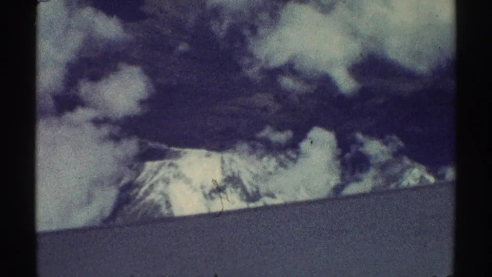 Faded 8mm home-movie frame of distant snow-capped mountains and clouds seen from an airplane during 1969 travel footage.