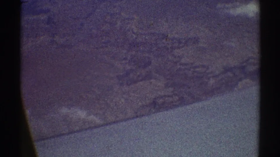 8mm frame from a 1969 flight showing arid ridges and pale land beyond a dark airplane window divider.