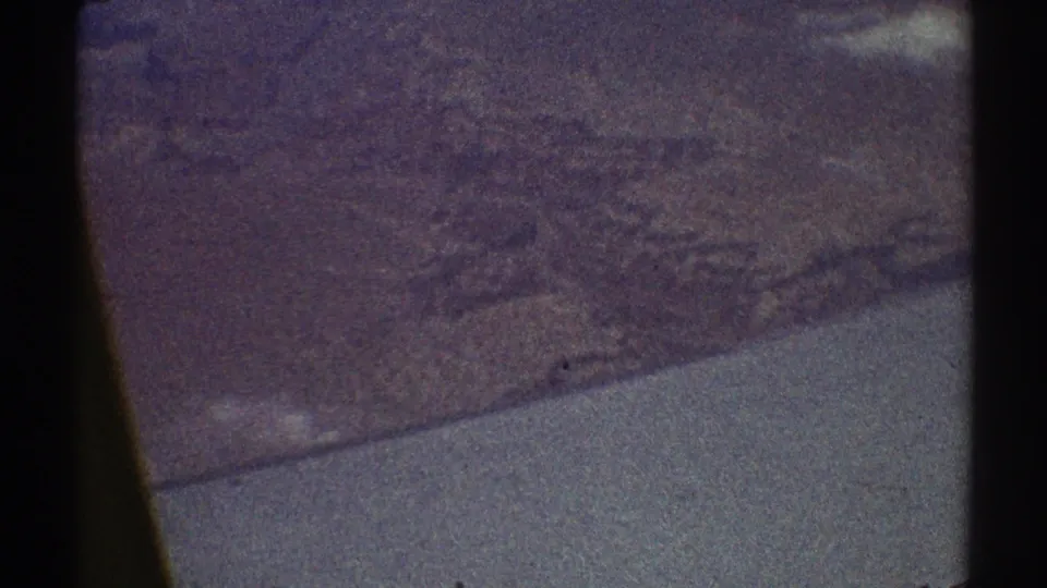 1969 8mm aerial home-movie view with soft, grainy landforms and a pale slanted aircraft edge or reflection at the bottom.