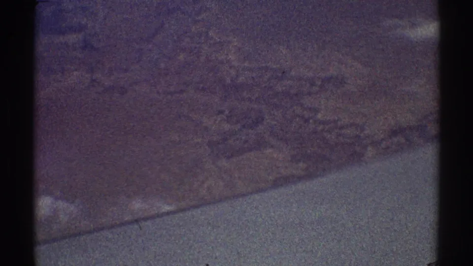 1969 8mm frame showing an indistinct aerial view over land or water, with dark vertical borders and muted purplish color.