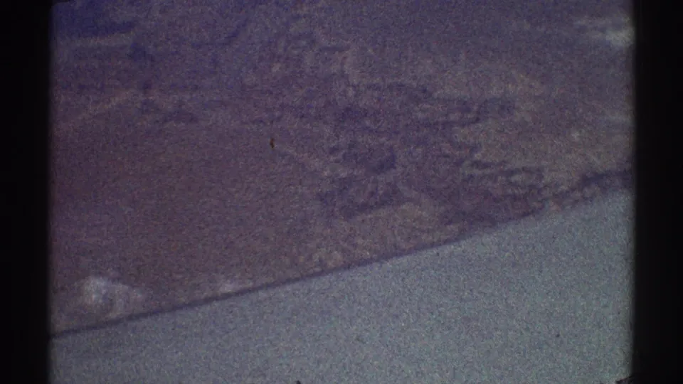 Vintage 8mm aerial frame with mottled land texture and a pale diagonal band that may be water, cloud, or reflected light.