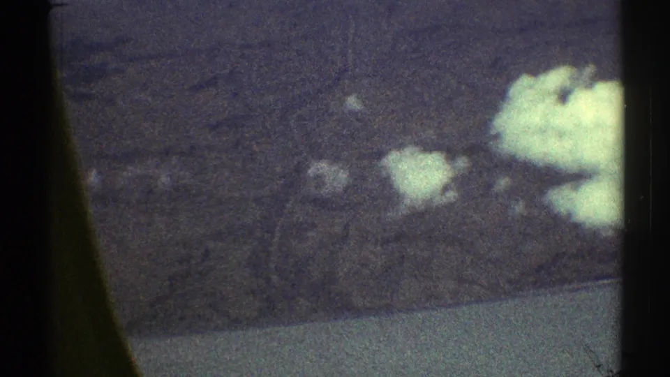 1969 8mm in-flight view with small white clouds over dark terrain, framed by the airplane-window edge.