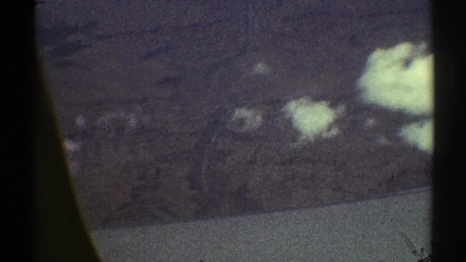 1969 8mm airplane-window footage showing bright clouds over dark terrain with a curved dark window edge at left.