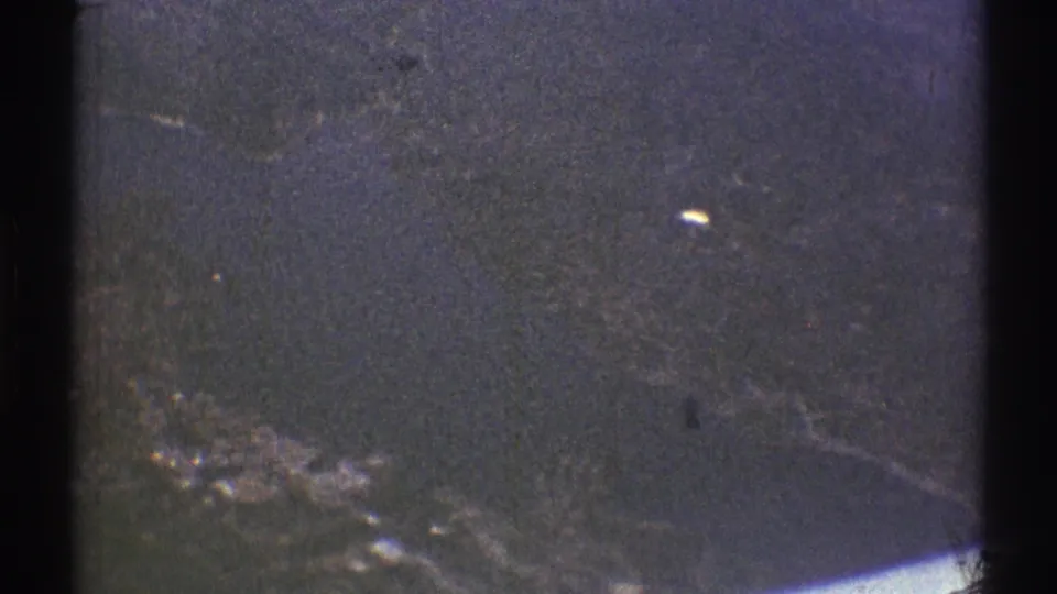 Aircraft-window view from the 1969 home movie, showing dark land or water below with scattered pale highlights and heavy 8mm grain.