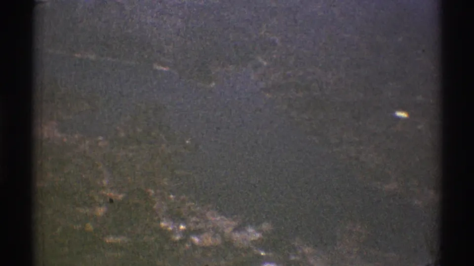 1969 8mm aerial frame showing indistinct terrain or water below, with bright patches and a dark aircraft-window border along the side.