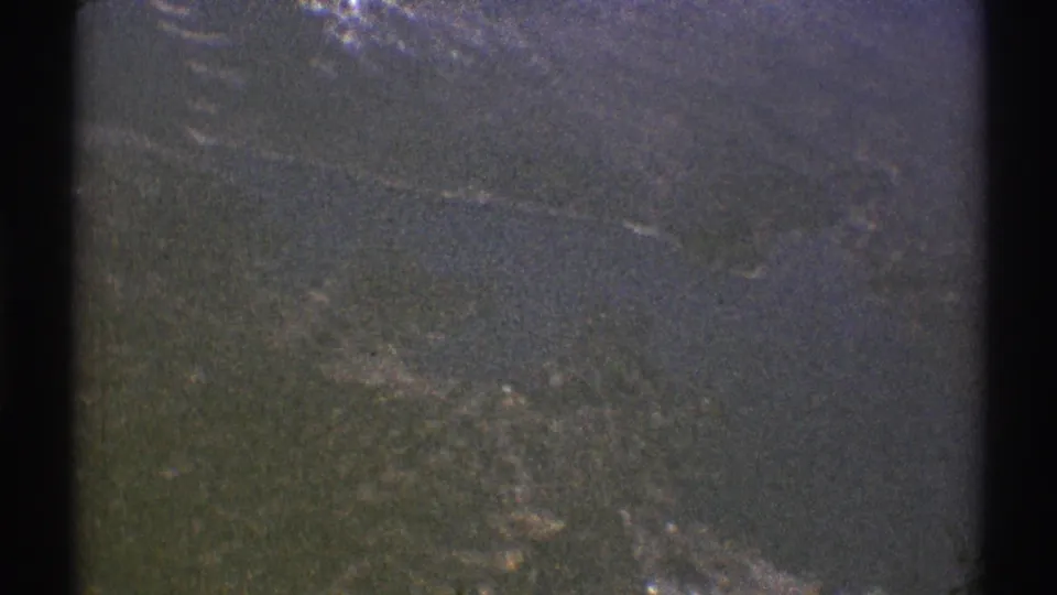 Late-1960s 8mm frame from an aircraft-window view, with dark land or water, scattered reflections, and visible edge shadowing from the film scan.