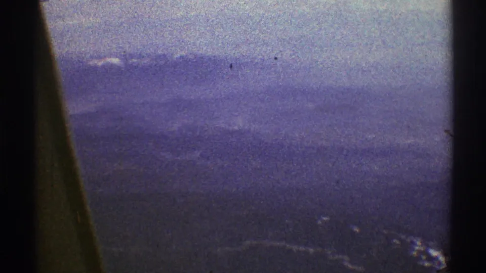 1969 8mm aerial view near Los Angeles with layered hazy ridges, scattered snow, and dark window framing in the foreground.