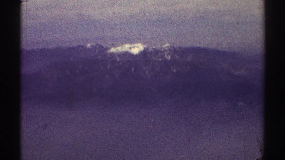 1969 8mm Los Angeles-area landscape frame with a snow-covered ridge beyond hazy hills and muted foreground fields.