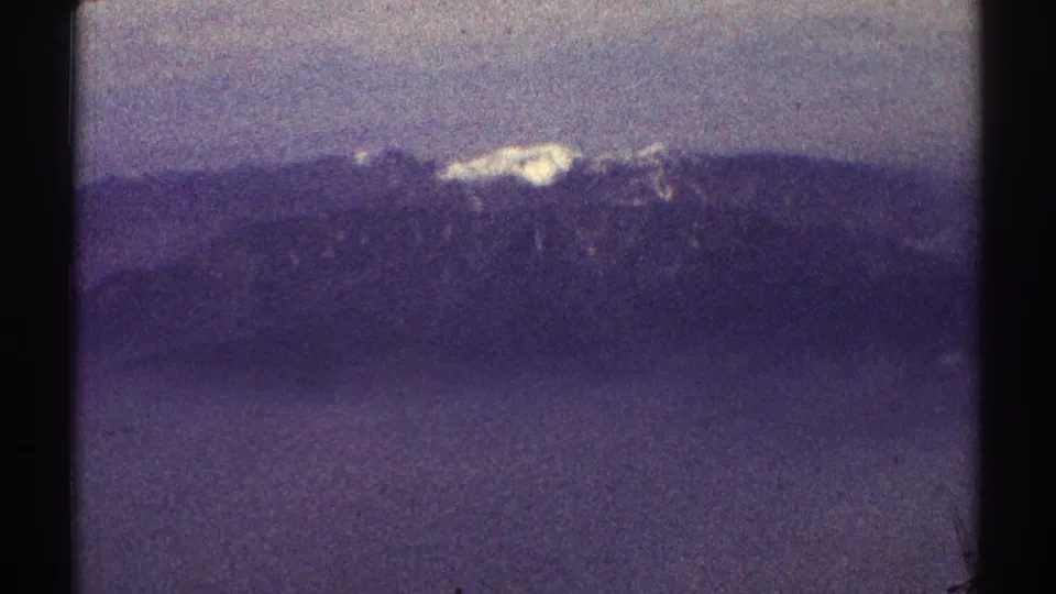 1969 8mm frame showing a distant snowy mountain ridge, hazy lower slopes, and visible home-movie film texture.