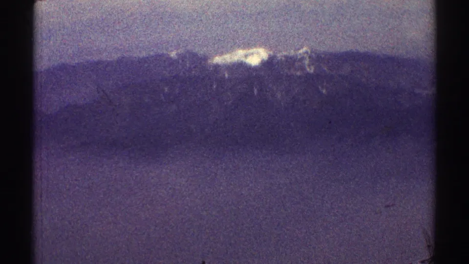 1969 Los Angeles-area 8mm frame showing a distant snow-covered summit beyond dark mountain ridges in a hazy purple sky.