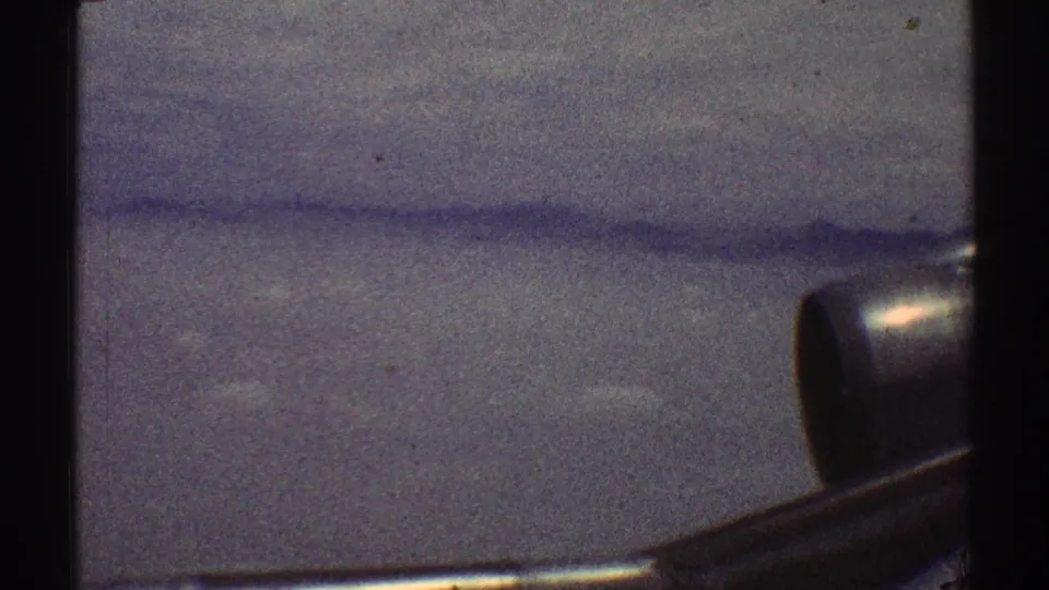 1969 8mm home-movie view from an airplane window, showing hazy clouds below and a partial aircraft wing or engine area along the right side.