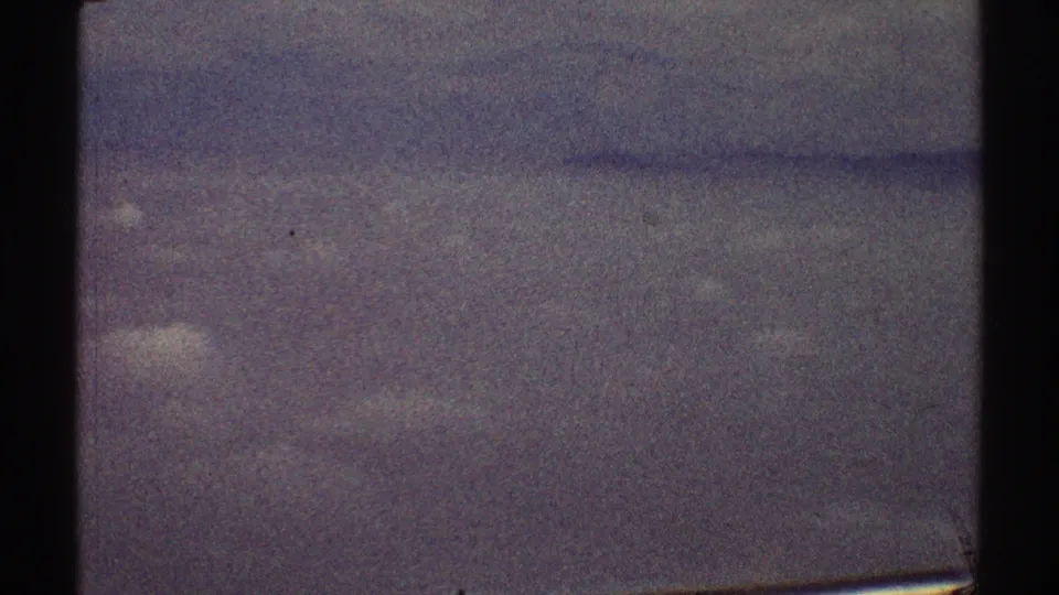 The 1969 home-movie frame shows a soft, grainy cloud layer through an airplane window, with dark frame edges and no identifiable ground features.