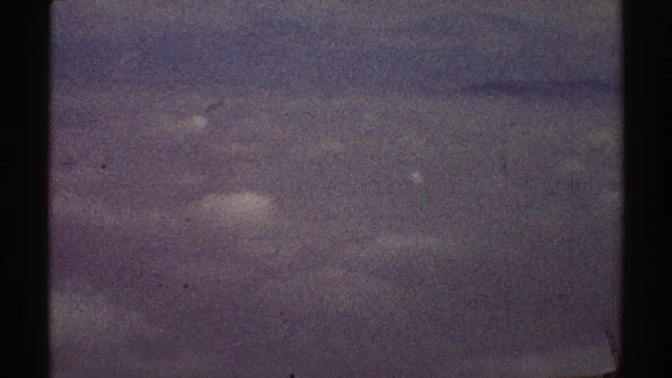 A muted 8mm airplane-window frame shows indistinct clouds below, a dark vertical window divider, and no visible people or landmarks.