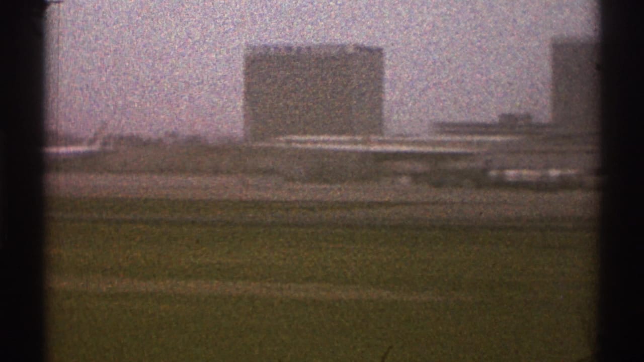 Grassy runway edge with distant airport buildings and parked aircraft beyond dark vertical frame edges.