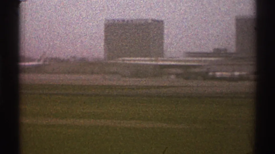 1969 Los Angeles airport view from a moving vehicle, with grass in the foreground and distant buildings and aircraft under a hazy sky.