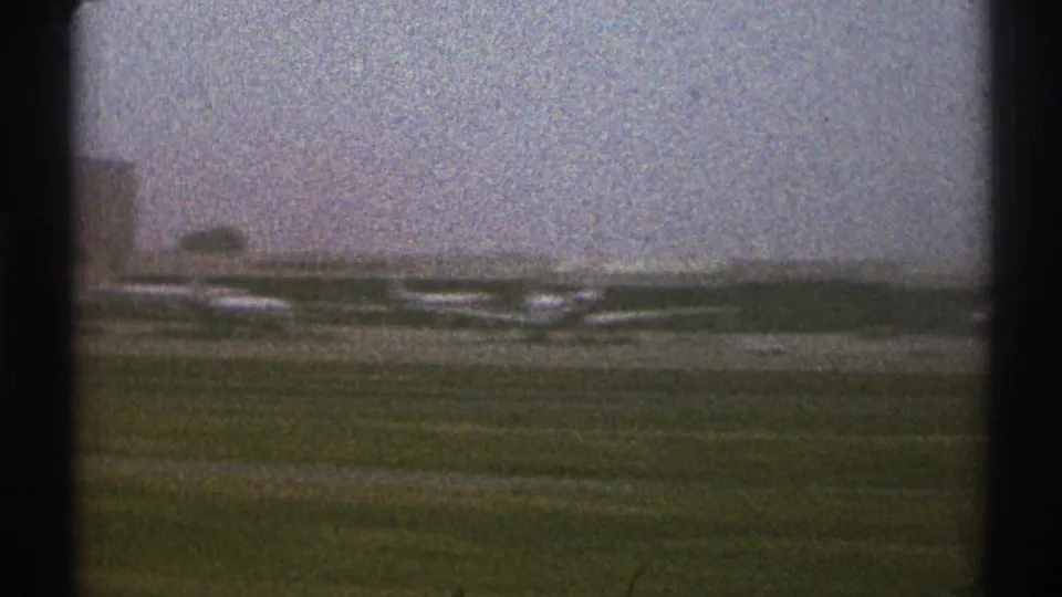 Grainy 8mm side view of the Los Angeles airport area, with parked planes and low buildings seen beyond a grassy strip.