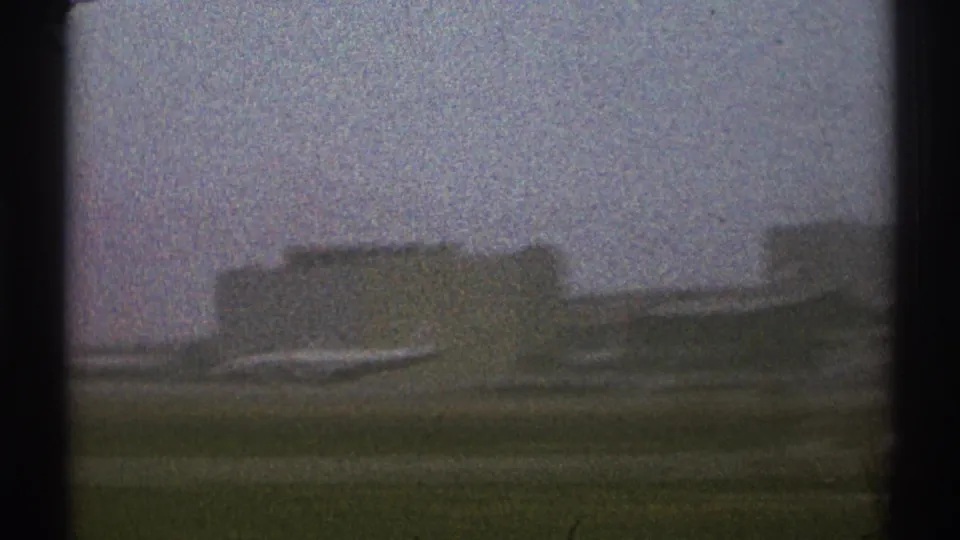 Moving 8mm view across a grassy airport edge toward distant planes and blocky terminal or hangar buildings in Los Angeles.