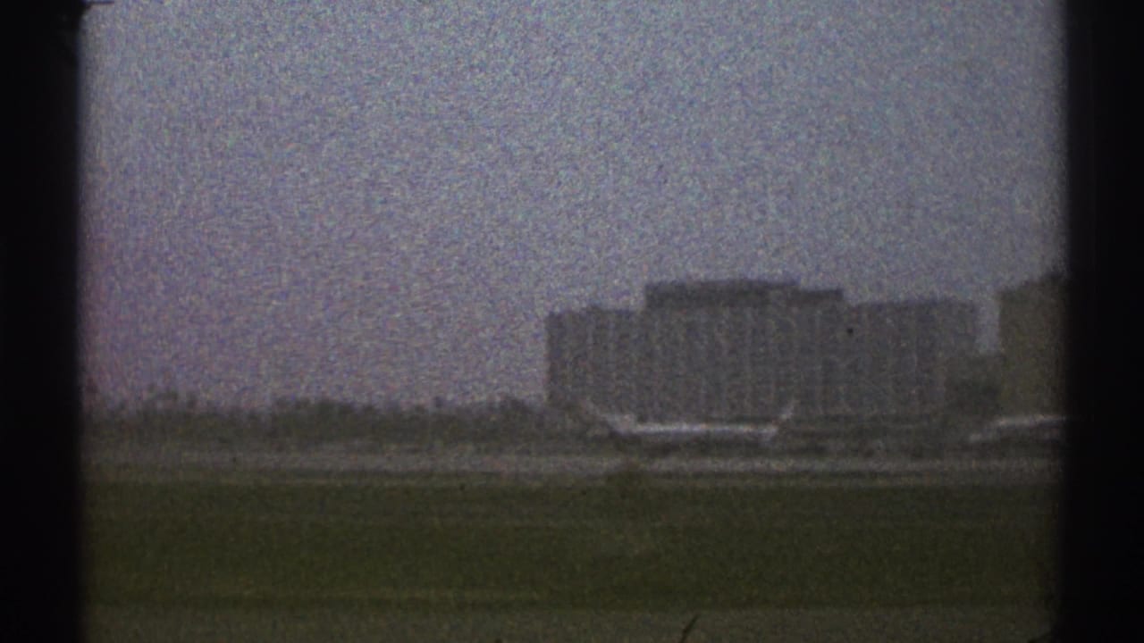 Distant airport building beyond grass, seen through dark window edges under an overcast sky.
