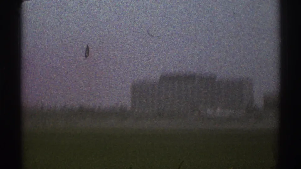 A grainy 1969 Los Angeles airport-area frame shows a low building beyond open grass, viewed from a moving car.