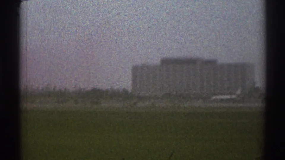 The moving 8mm view catches low airport structures on the horizon beyond a green field in Los Angeles in 1969.