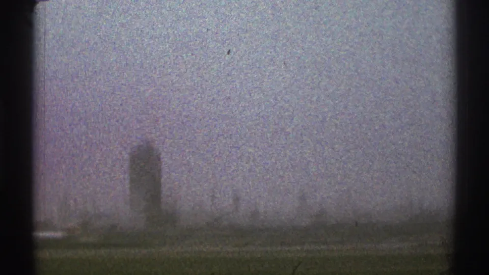 1969 Los Angeles-area 8mm frame showing a hazy distant airport control tower and low tarmac structures from a vehicle viewpoint.
