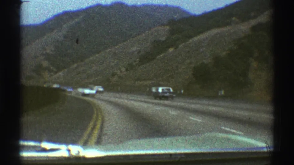 1969 Los Angeles-area road footage showing a curving highway, dry hills, oncoming cars, and the edge of the filming vehicle hood.