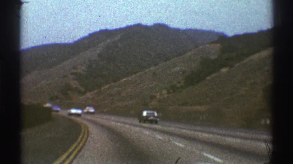 1969 Los Angeles-area 8mm frame of highway traffic passing through dry rolling hills beneath a pale sky.