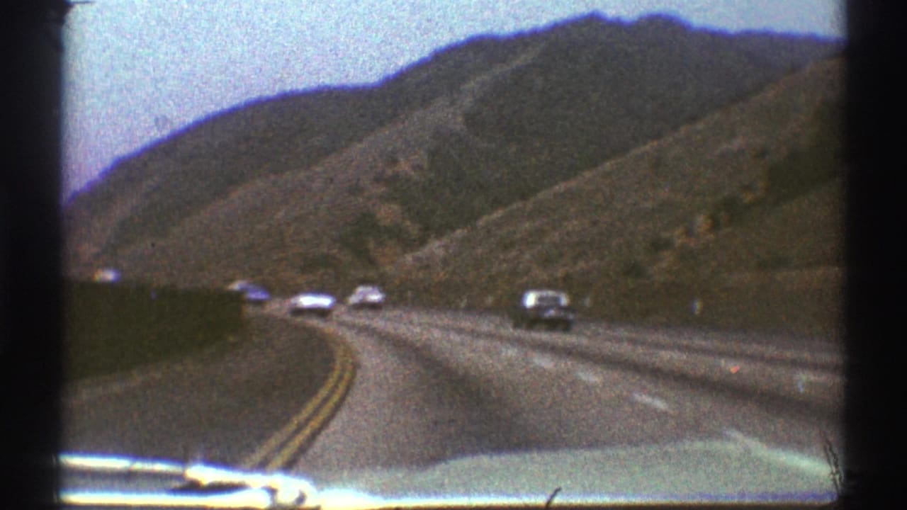 1969 Los Angeles-area home-movie view from inside a car on a winding road bordered by dry hills and passing traffic.