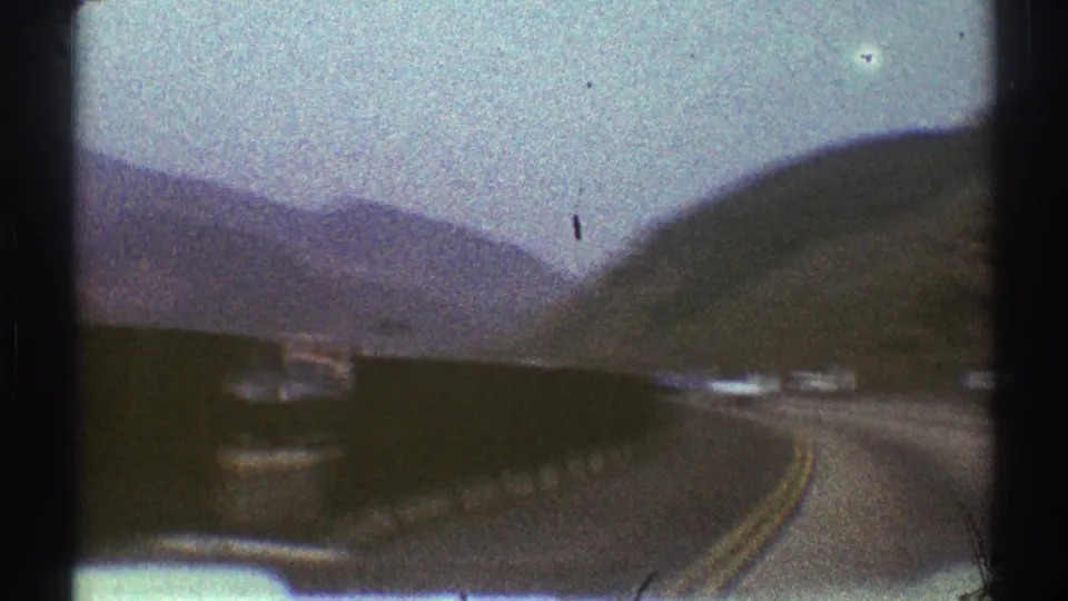Passenger-side view of a curved Los Angeles-area mountain roadway in 1969, with a barrier wall, distant cars, and dry slopes.