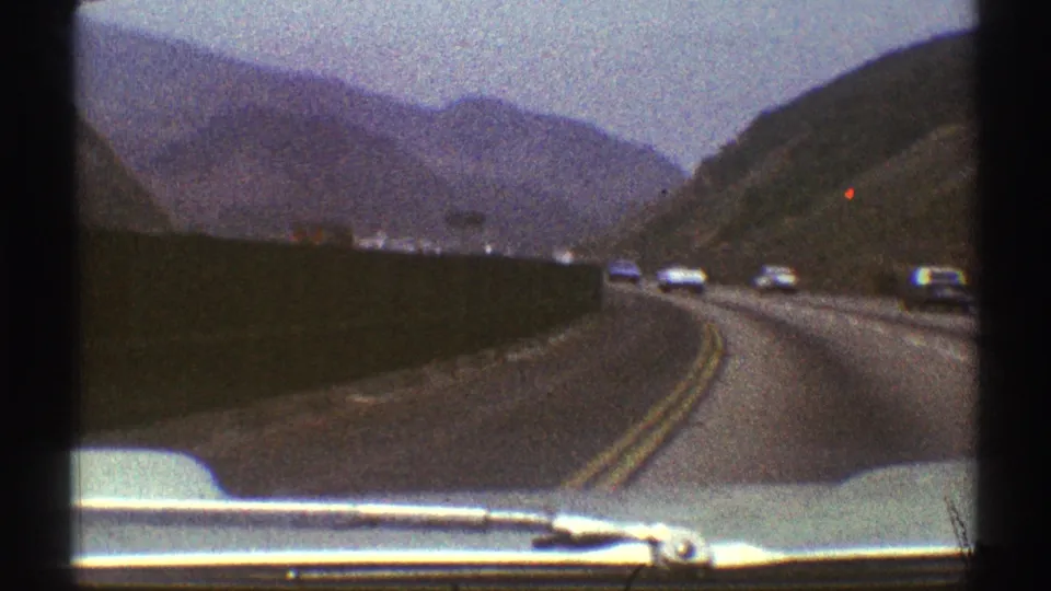 1969 home-movie windshield view of cars on a winding Los Angeles-area road with dry hillsides and distant mountains.