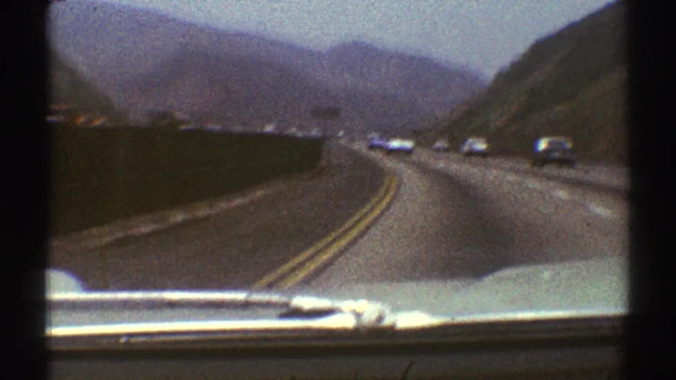 Forward-facing 1969 Los Angeles-area driving view showing a curving road, light traffic, dry hills, and the filming car's hood.