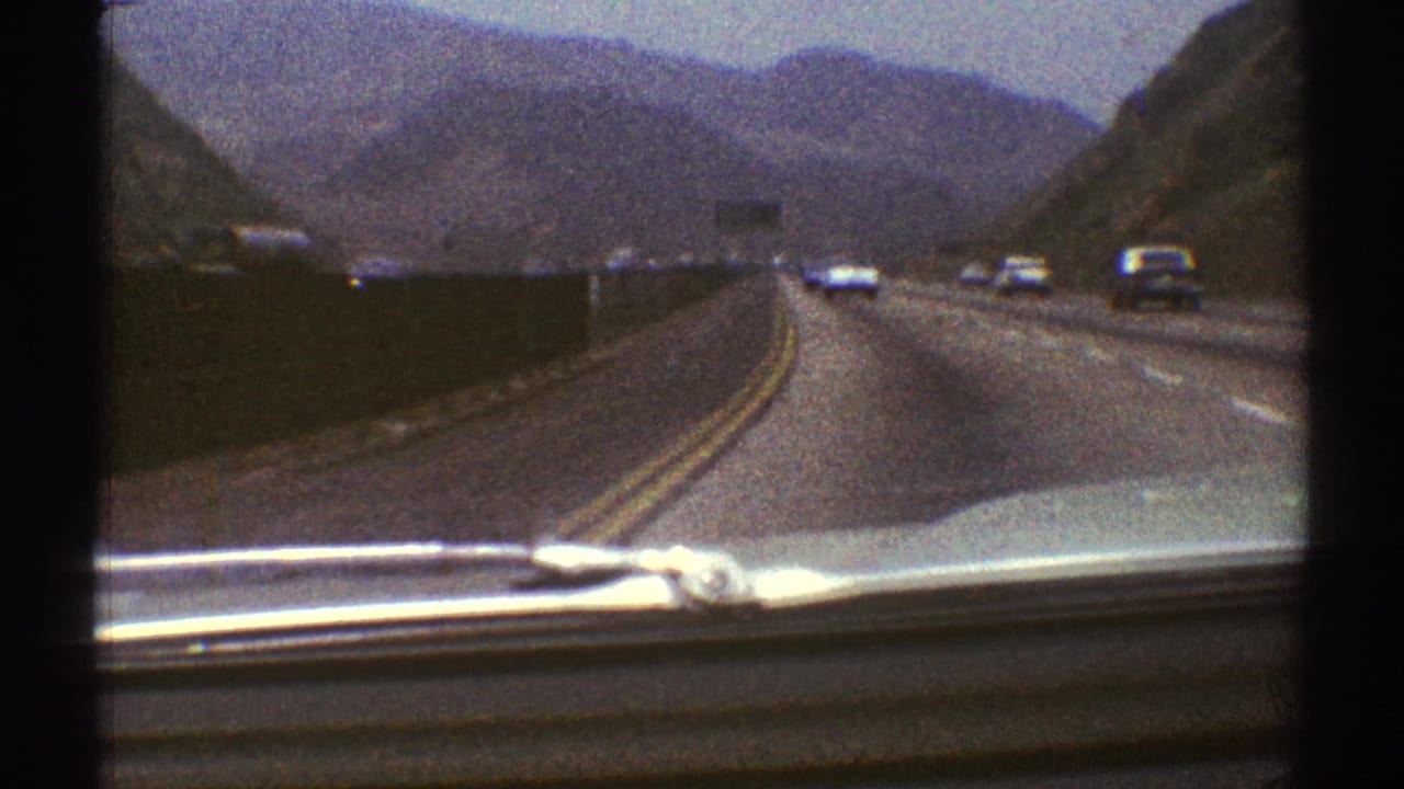 1969 Los Angeles-area 8mm windshield view of cars traveling on a multi-lane mountain highway under cloudy sky.