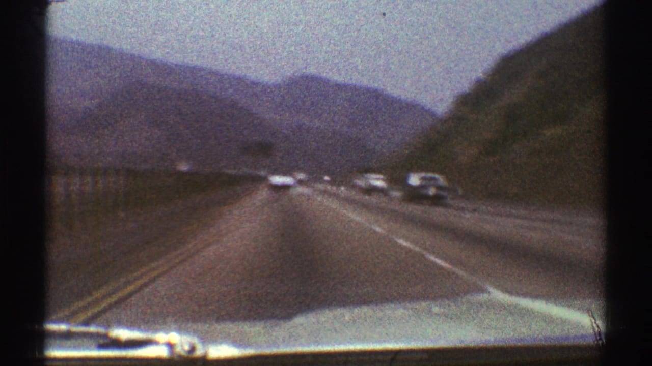 Forward view over a car hood on a highway bordered by hills and distant mountains.