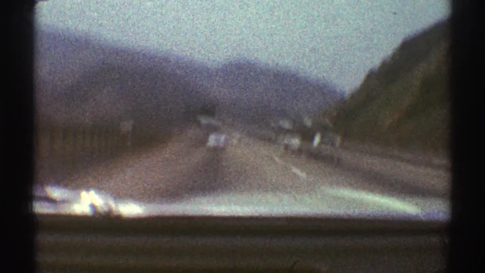 Soft, grainy 8mm frame from a moving car on a mountain highway, with road lanes, traffic, and hillside visible.