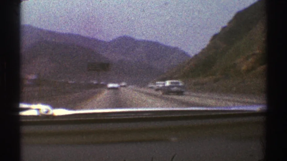 1969 8mm windshield view of cars on a Los Angeles-area mountain highway, framed by dark film edges and cloudy sky.